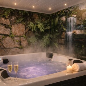 A hot tub with two glasses of champagne and a candle, surrounded by a waterfall mural and greenery.