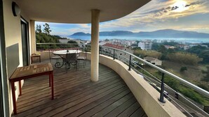 A balcony with a wooden table and chairs, overlooking a coastal cityscape with buildings and mountains. - Appartement Sole - Vue Splendide sur le Golfe de Porto-vecchio (Porto-Vecchio)