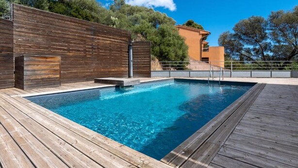 A rectangular swimming pool with a wooden deck surrounding it, a metal ladder, and a wooden fence. - Appartement Sole - Vue Splendide sur le Golfe de Porto-vecchio (Porto-Vecchio)