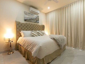 A bedroom with a tufted headboard, a bed with white bedding, a bedside lamp, and a large window with curtains.