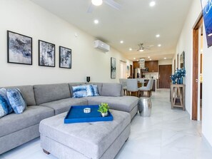 A modern living room with a grey sectional sofa, a blue coffee table, and a small plant. There is a dining area in the background with a kitchen visible.