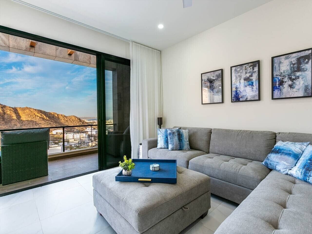 Ein modernes Wohnzimmer mit einem grauen Sofa, einem blauen Tablett und Blick auf eine Berglandschaft durch ein großes Fenster.