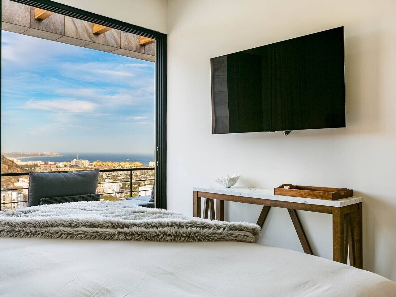 A bedroom with a large bed, a wooden nightstand, a flat-screen TV mounted on the wall, and a view of the city through a large window.