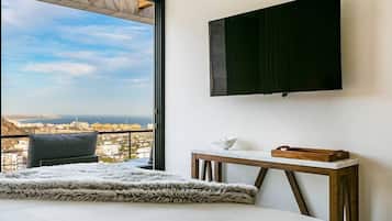 A bedroom with a large bed, a wooden nightstand, a flat-screen TV mounted on the wall, and a view of the city through a large window.