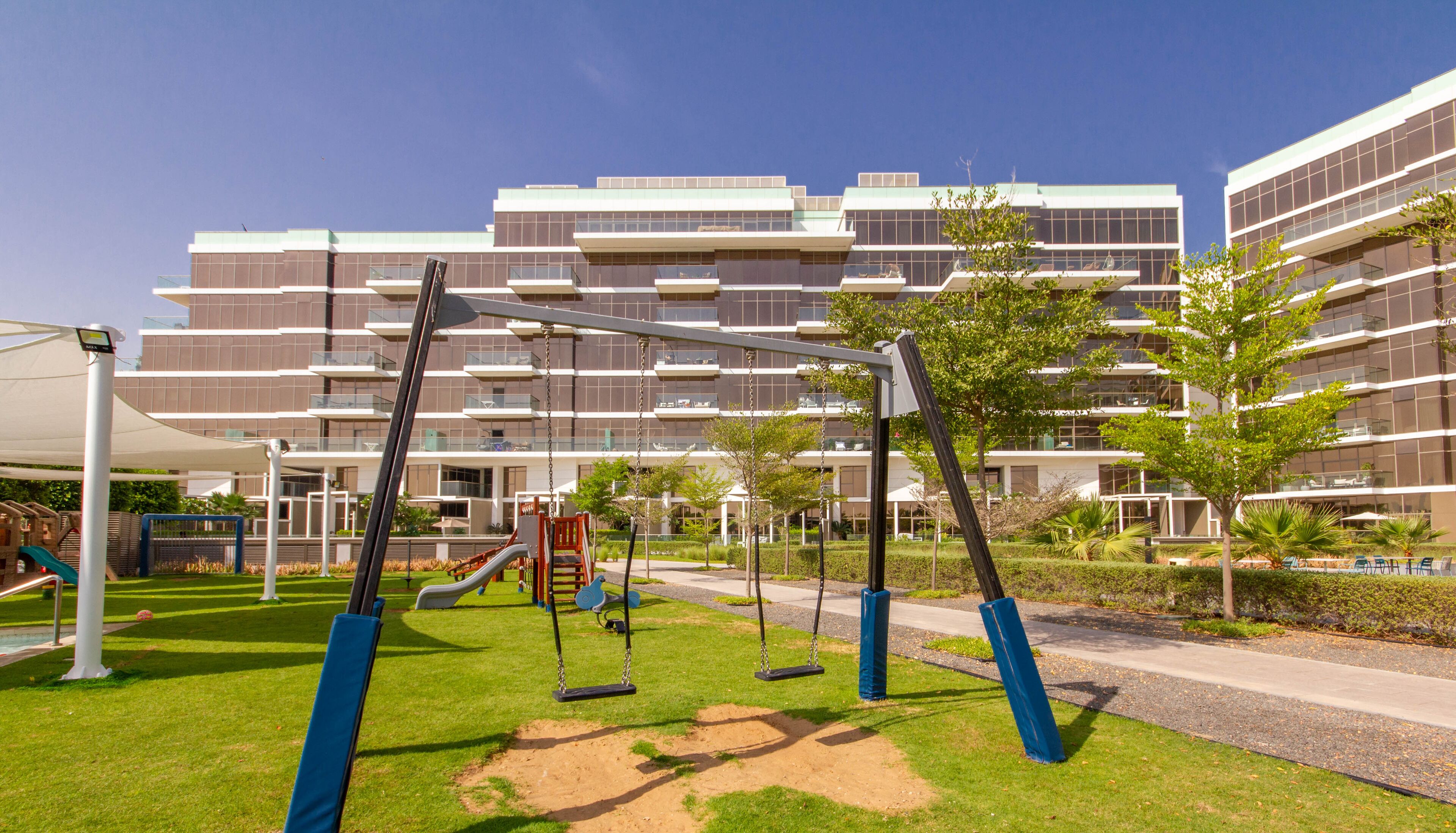 Un parque infantil con columpios y toboganes, rodeado de césped cuidado y árboles, con edificios de apartamentos modernos al fondo.