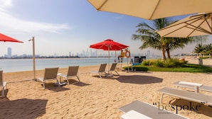 A beachside seating area with sun loungers and umbrellas, overlooking a body of water with a city skyline in the distance. - Bespoke Residences - Th8 Palm Jumeirah (Dubai)