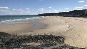 Une vaste plage de sable fin aux vagues douces, un premier plan rocheux et, au loin, une côte bordée de bâtiments et de verdure.