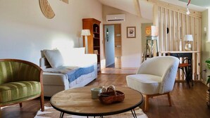 A cozy living room with a sofa, armchair, and coffee table, wooden beams on the ceiling, and a bookshelf.