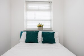 Un dormitorio con una cama, paredes blancas, un ventanal con persianas y una pequeña planta en maceta sobre el alféizar.