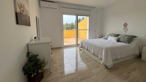 Ein Schlafzimmer mit einem Bett, einem Schreibtisch, einer Zimmerpflanze und einer Glastür, die auf einen Balkon führt.