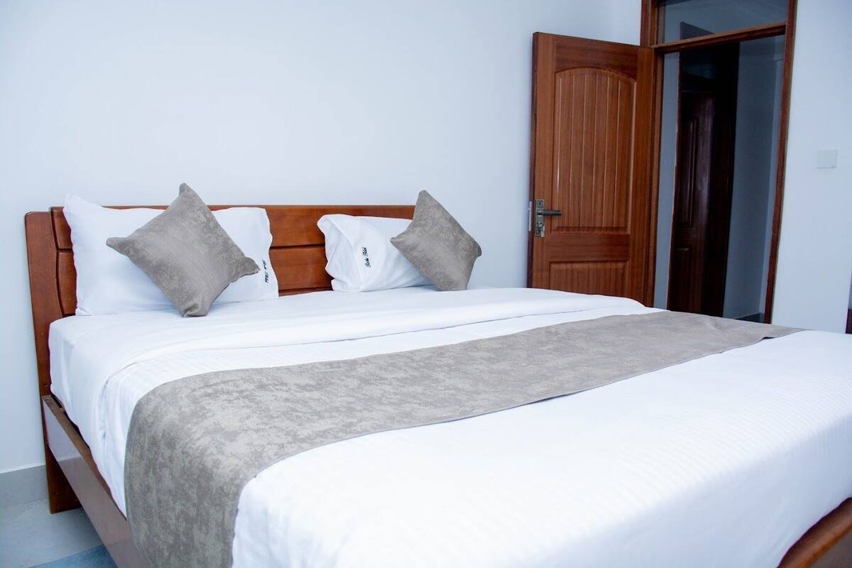 Cama bem arrumada com roupa de cama branca e manta cinza, cabeceira de madeira e dois travesseiros cinzas.