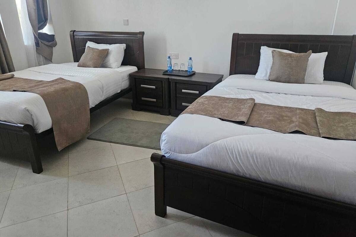 Quarto de hotel com duas camas, cada uma com cabeceira de madeira, roupa de cama branca e mantas marrons. Há uma pequena mesa de cabeceira com uma garrafa de água e copos.