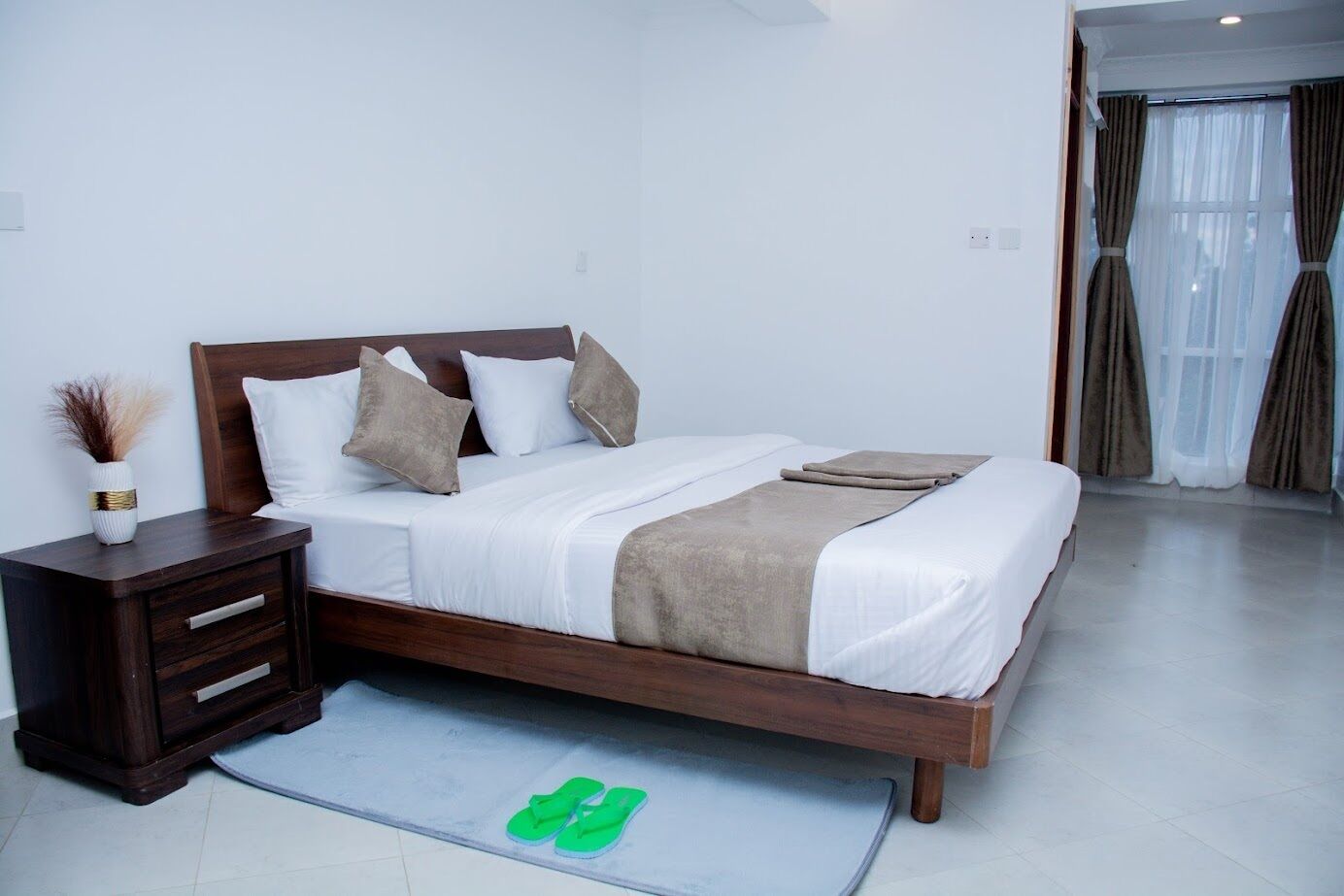 Un dormitorio ordenado con cama de madera, ropa de cama blanca, una mesita de noche con cajones y un par de pantuflas verdes en el suelo.