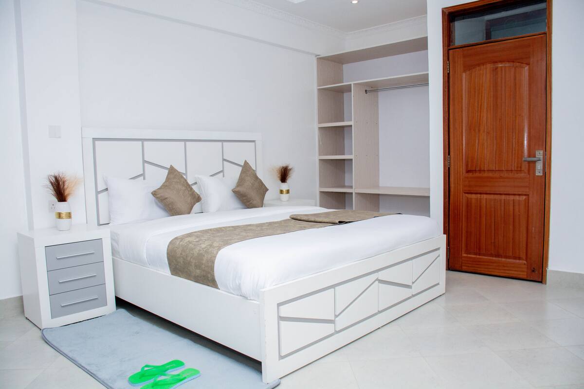 Quarto bem arrumado com cama branca, porta de madeira e guarda-roupa embutido.