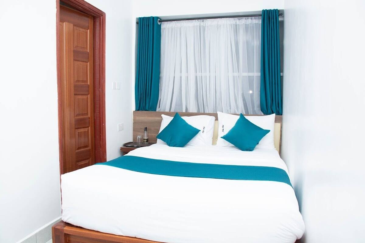 Quarto de hotel com uma cama, uma porta de madeira e uma janela com cortinas azul-turquesa.