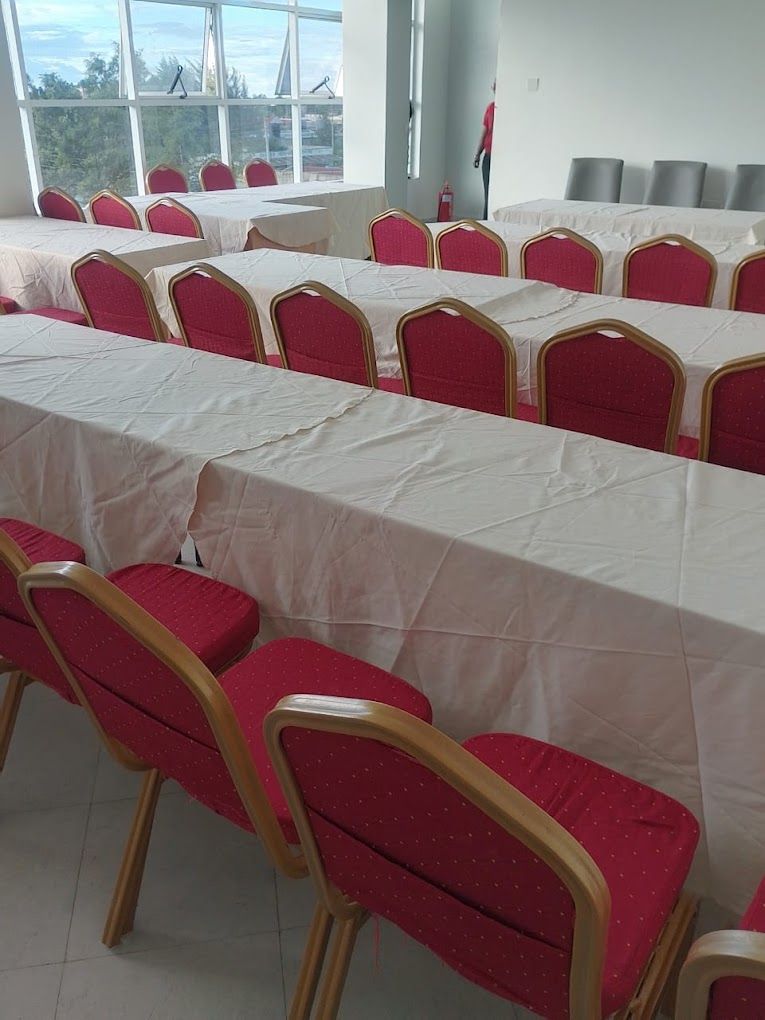 A conference room with rows of red chairs and white tables, large windows with a view of trees and buildings, and a white ceiling.