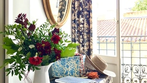 Una stanza con una valigia vintage, un vaso bianco con fiori, uno specchio e tende a motivi.