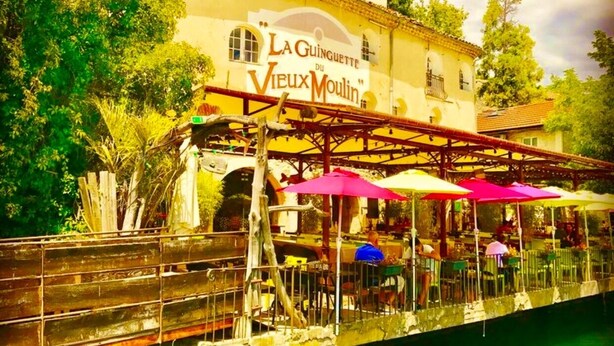 Un ristorante sul fiume con tavoli all'aperto e un cartello che riporta la scritta 'La Guinguette du Vieux Moulin'.