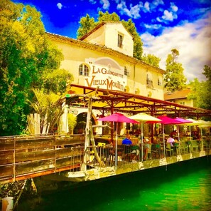 Un restaurant situé au bord de la rivière, doté d’une terrasse et d’une enseigne indiquant : « La Guinguette du Vieux Moulin ».
