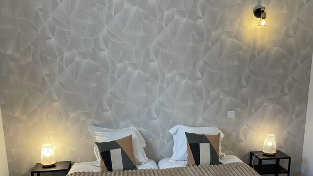 A hotel room with a textured geometric wall, a large bed with a beige quilt, two bedside lamps, and neatly folded towels on the bed. - Le Clos du Thoronet (Le Thoronet)