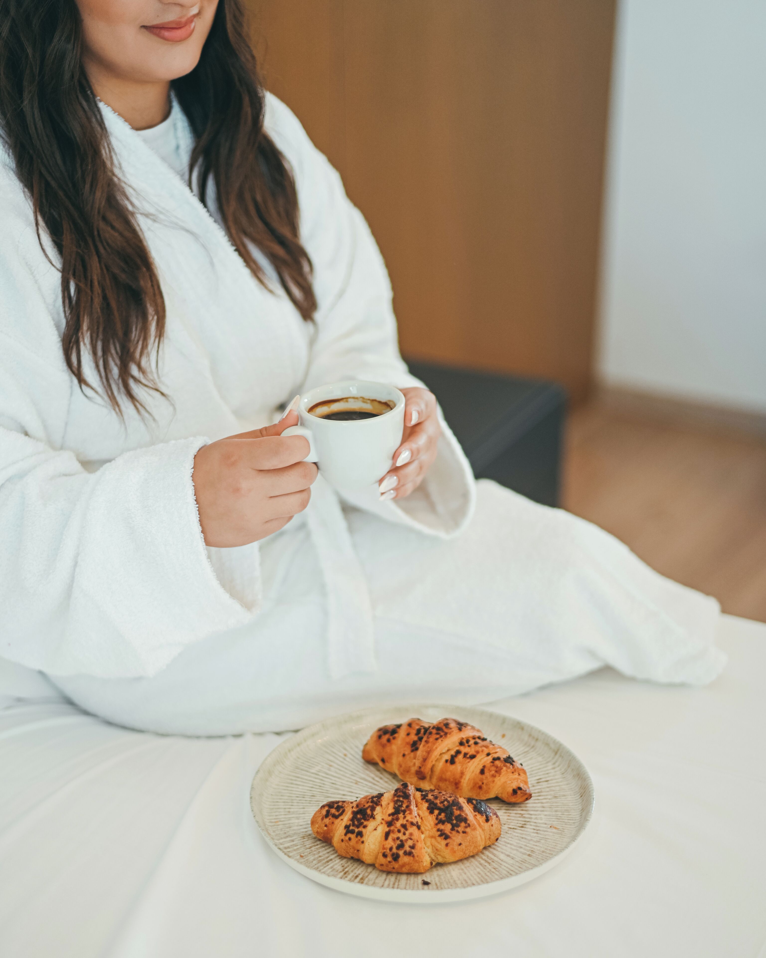 A person in a white robe holding a cup of coffee, sitting at a table with a plate of croissants.