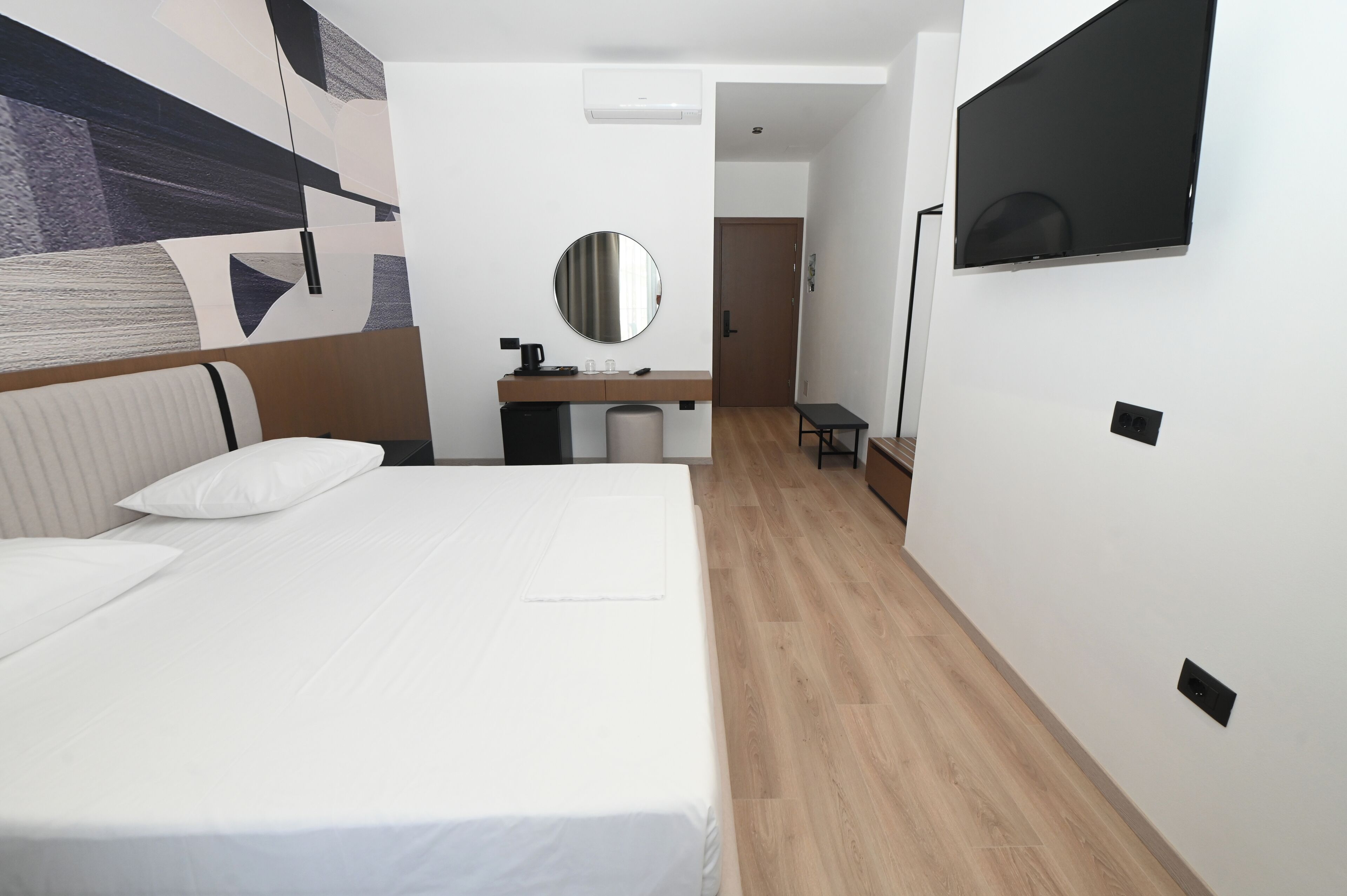 Habitación de hotel con una cama grande, una mesita de noche con espejo, un escritorio pequeño y un televisor montado en la pared.