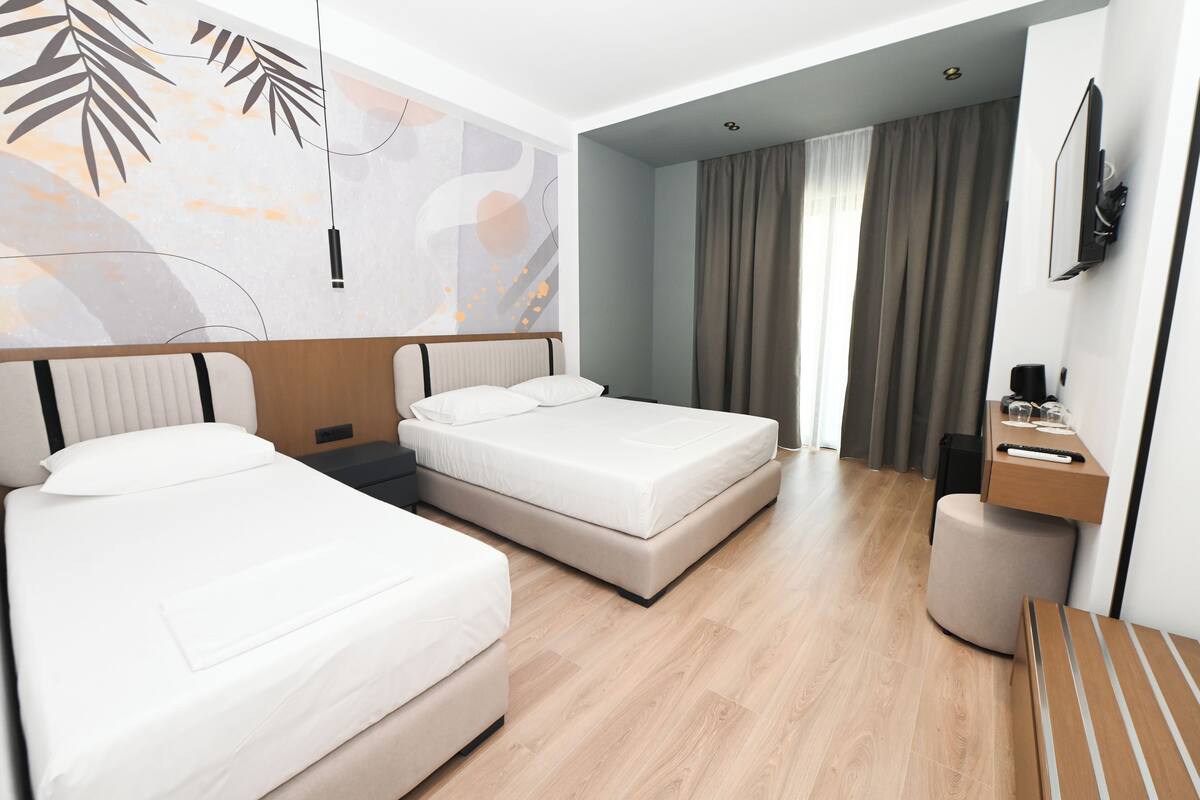 A hotel room with two beds, a desk, a chair, a TV, and a large window with curtains.