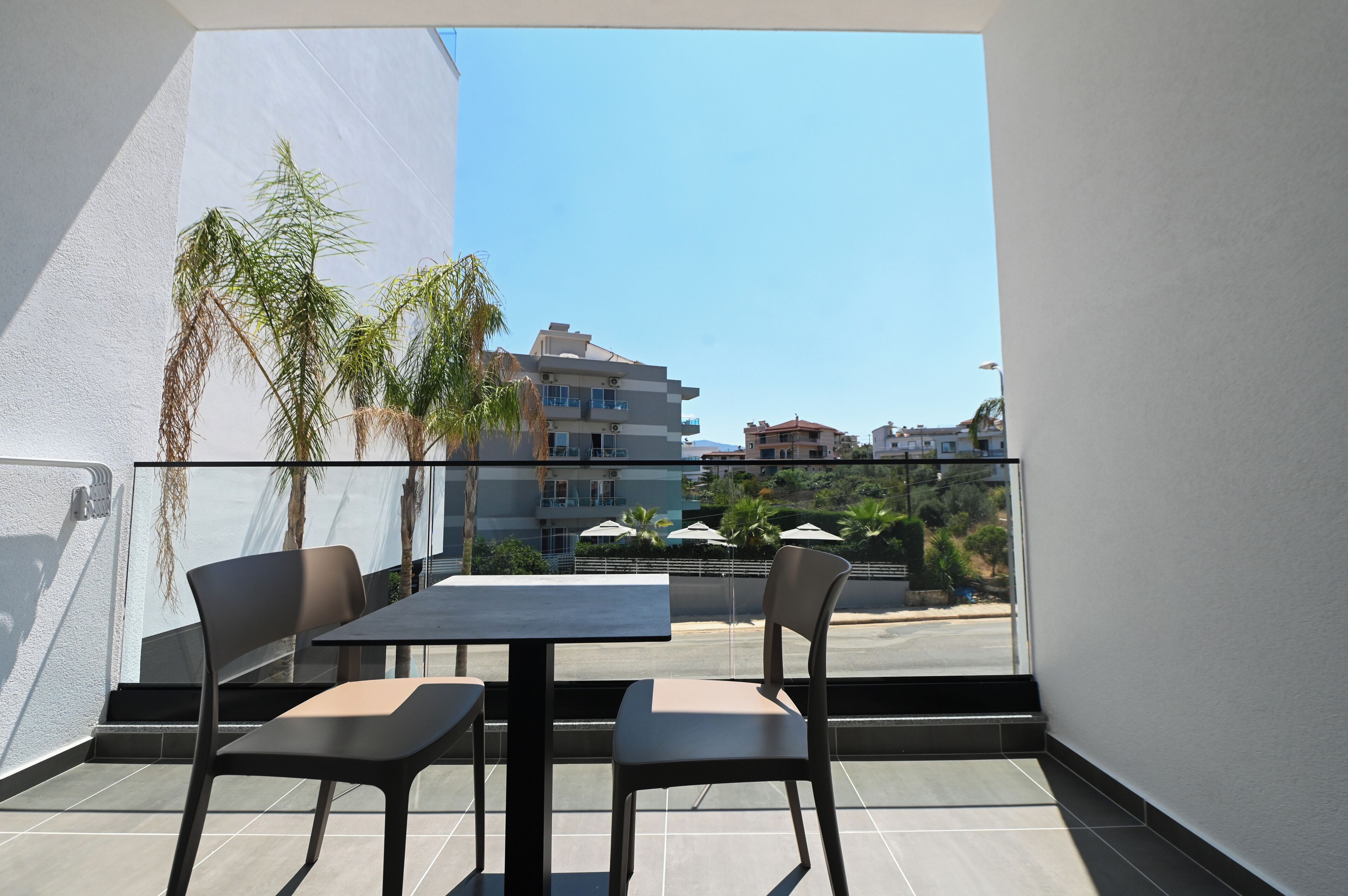 Un balcón con una mesa y sillas, con vista a una calle con edificios y palmeras.