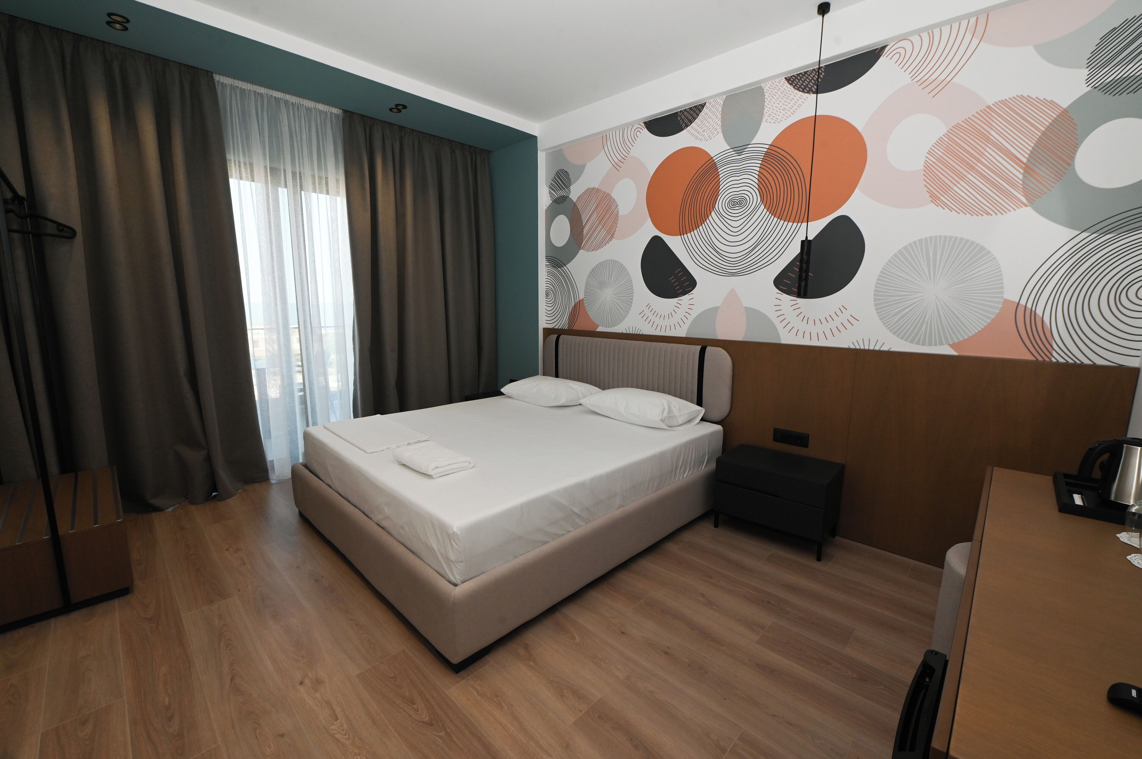 A modern hotel room with a large bed, wooden flooring, a nightstand, and a wall with abstract patterns.