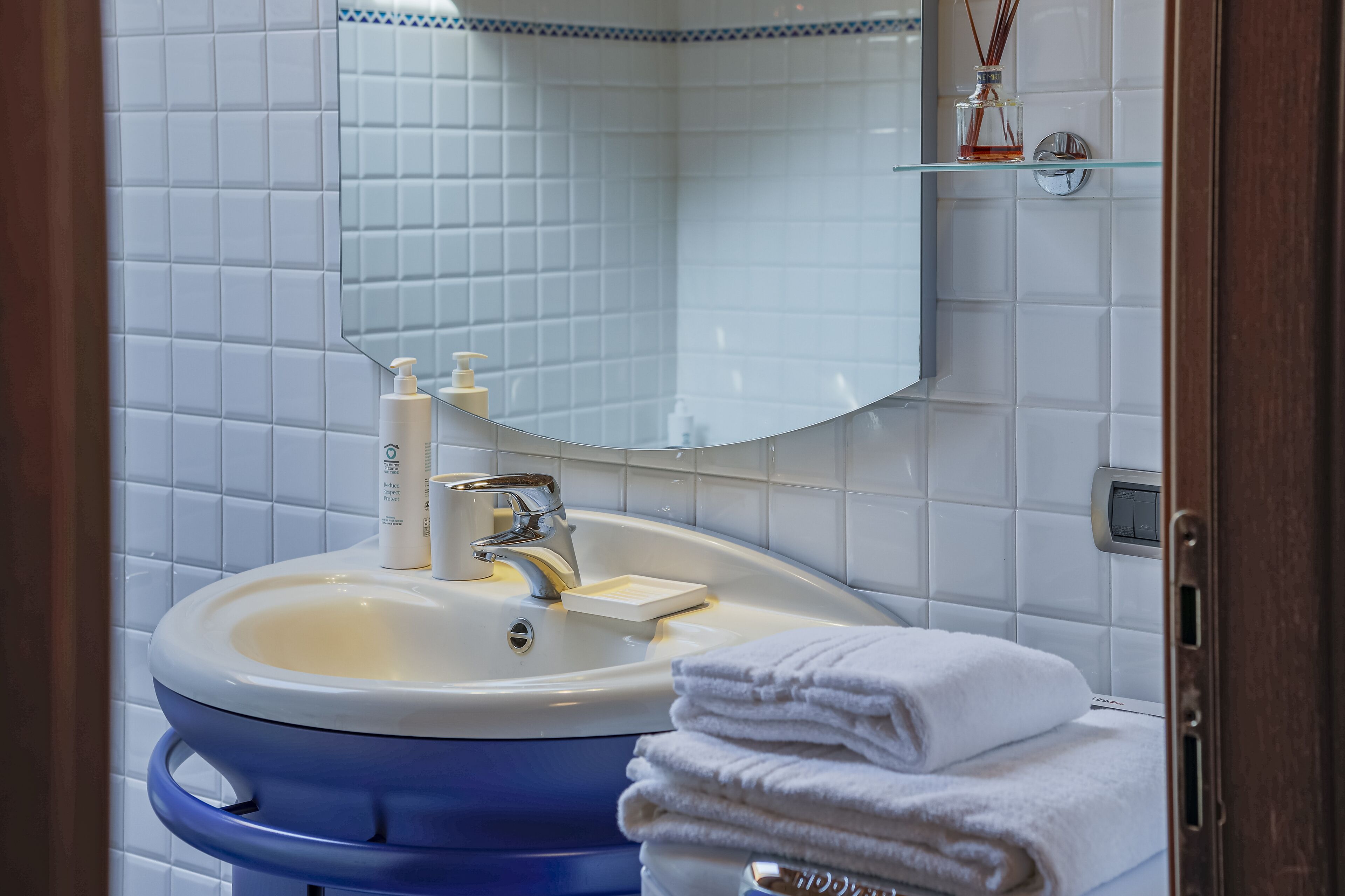 A bathroom with a white tiled wall, a blue sink, a mirror, and a glass shelf with toiletries.