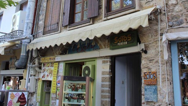 Un immeuble à plusieurs étages doté d’un balcon, avec un commerce au rez-de-chaussée et une enseigne indiquant « Sotos Pension ».