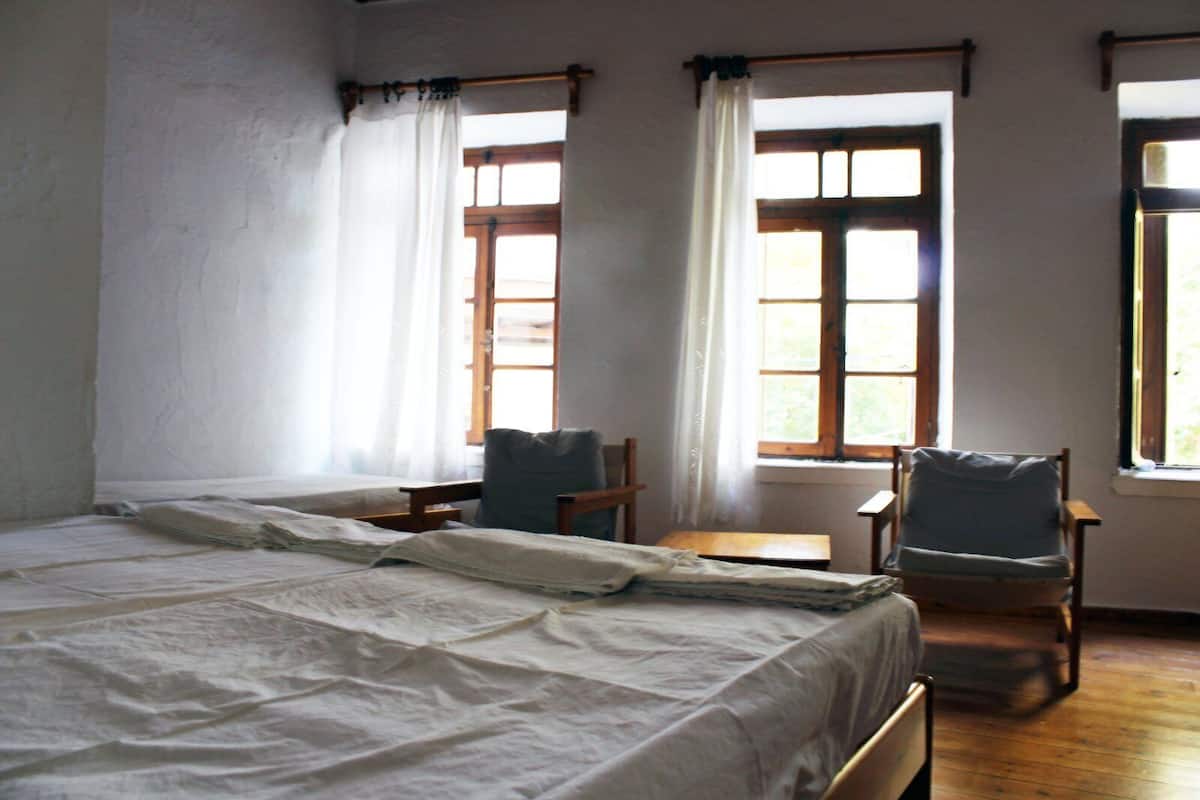 A room with a bed, two chairs, a small table, and windows with curtains.
