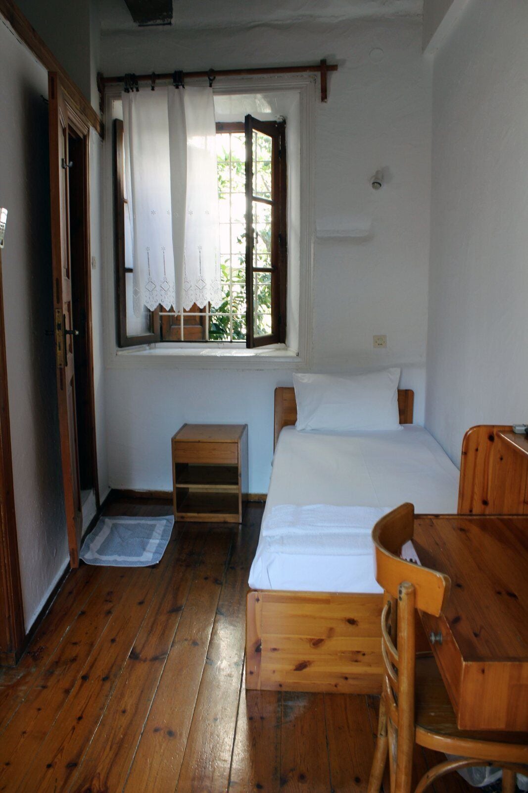 A small, simple room with a wooden floor, a single bed, a wooden desk, and a window with curtains.
