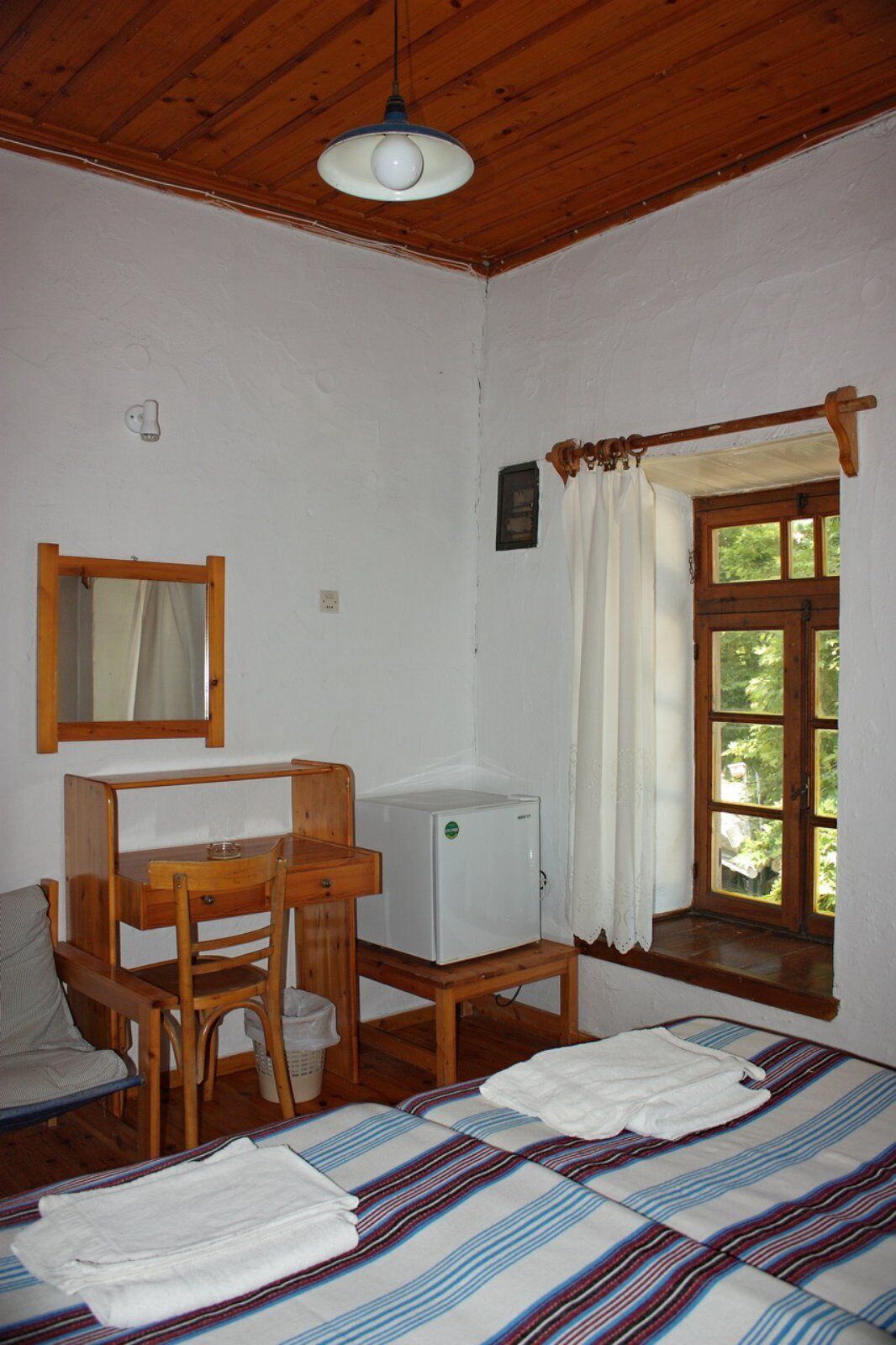 Una habitación pequeña y acogedora con cama, un escritorio de madera, un refrigerador pequeño y una ventana con cortinas.