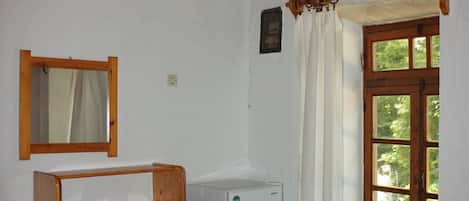A small, cozy room with a bed, a wooden desk, a small refrigerator, and a window with curtains.