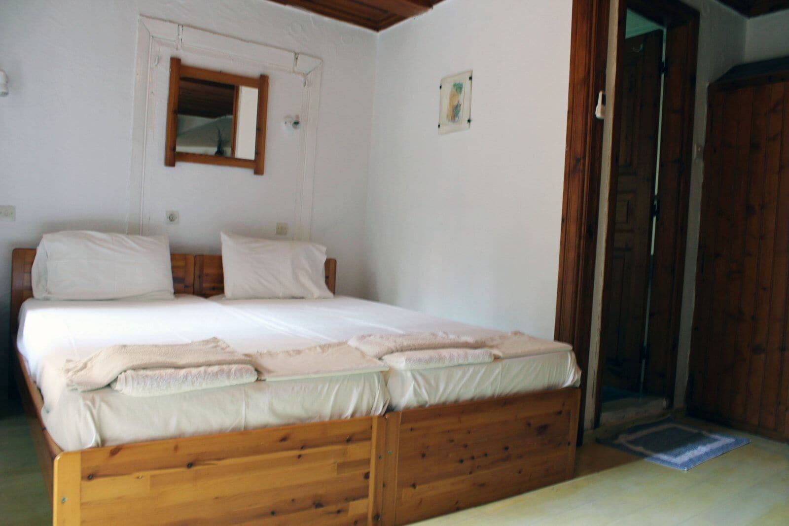Um quarto com uma cama de madeira, paredes brancas, uma porta de madeira e uma pequena janela.
