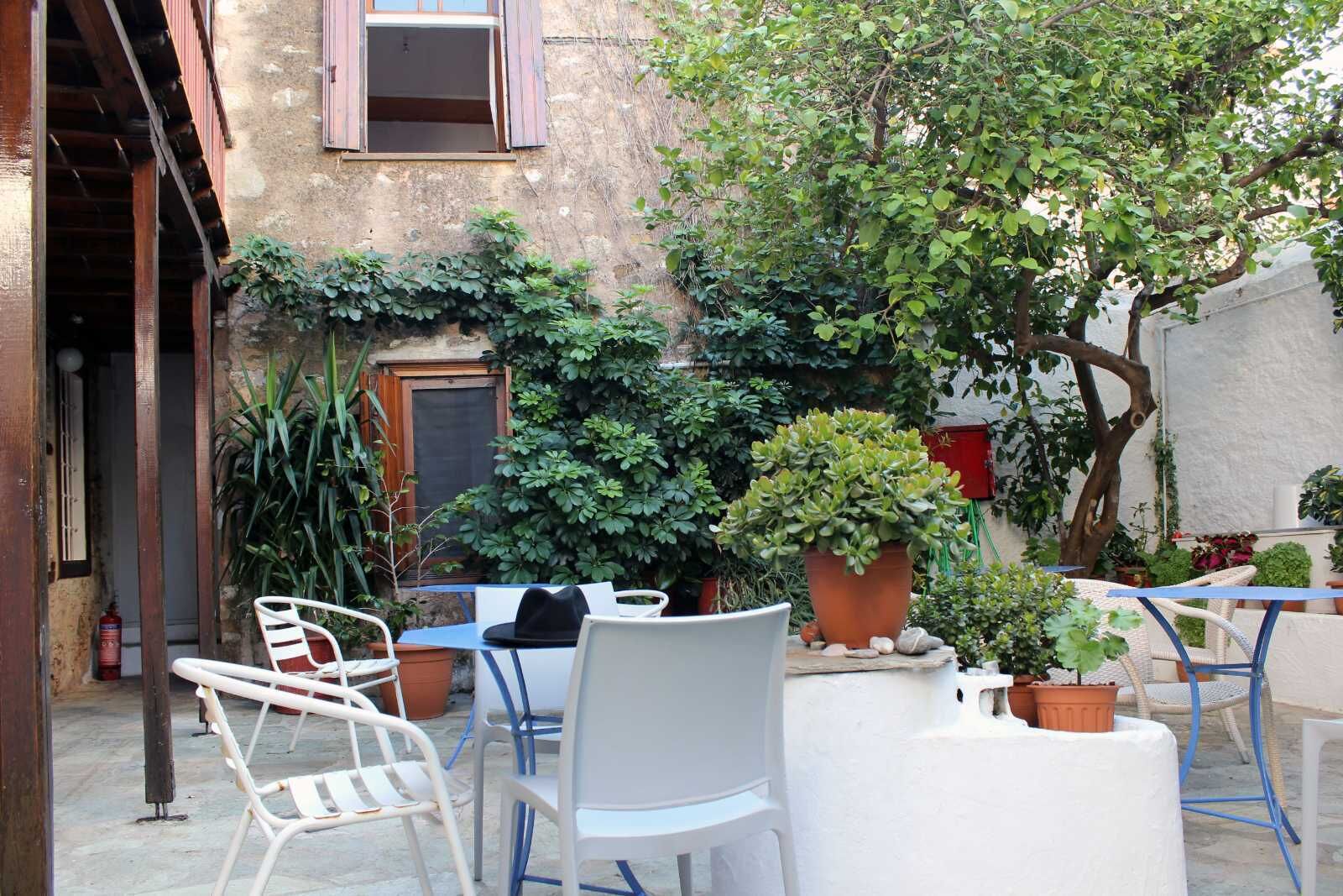 Un patio exterior con sillas y mesas de metal blanco, rodeado de macetas con plantas y un árbol.