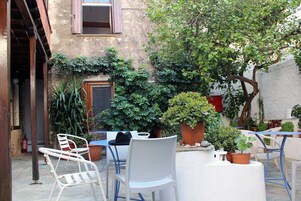 Un patio exterior con sillas y mesas de metal blanco, rodeado de macetas con plantas y un árbol.