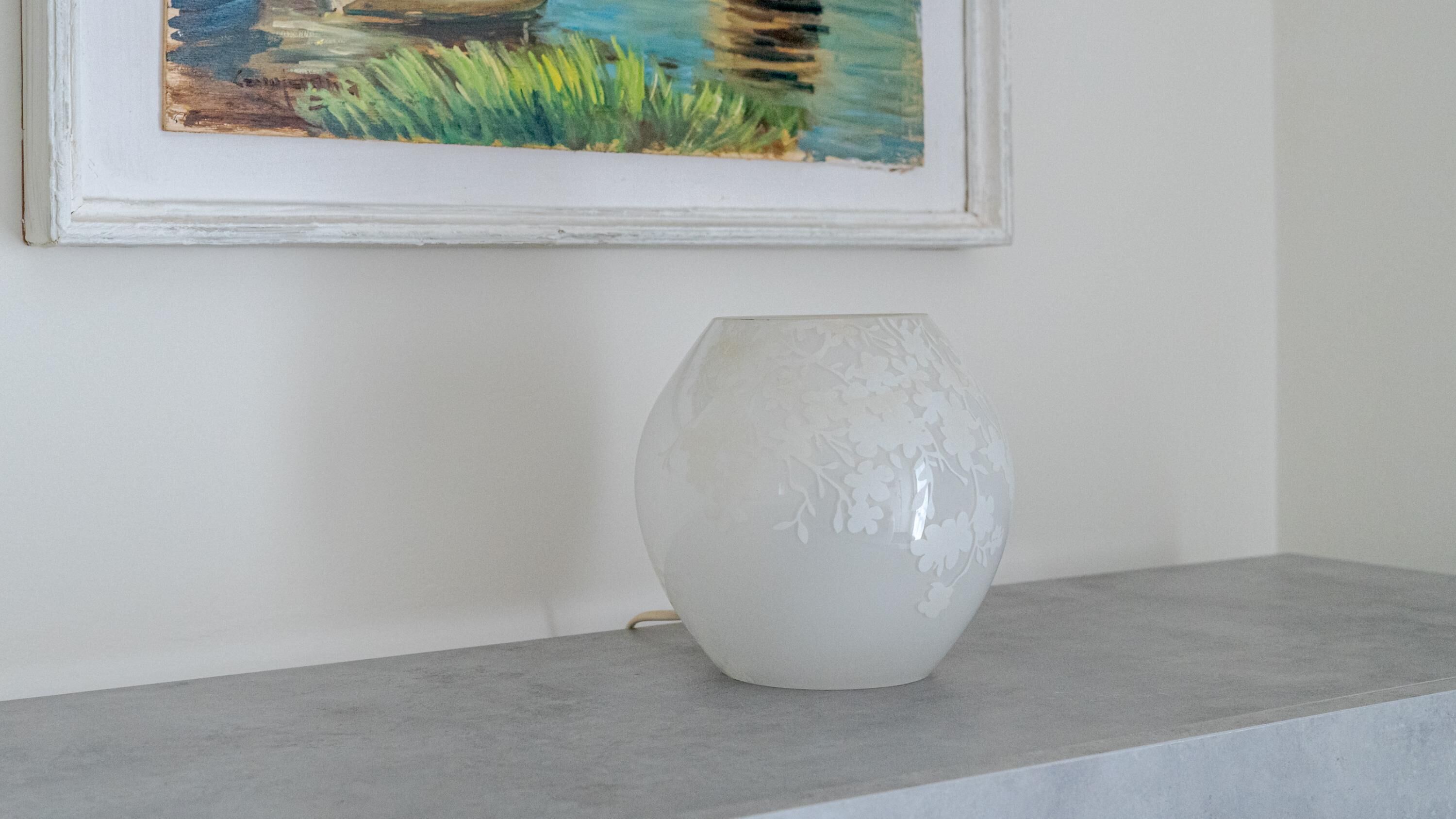 A white vase on a grey shelf against a white wall with a framed painting above it.