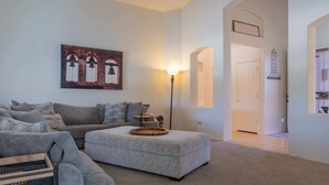 Un salon avec un canapé gris, un pouf gris et une lampe sur pied. Il y a un tableau au mur et une porte avec un porte-manteau.