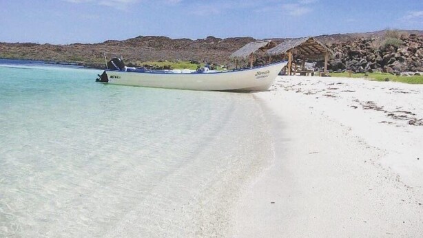 Ein Boot an einem sandigen Strand mit klarem Wasser, einem mit Stroh gedeckten Gebäude und hügeliger Küstenlandschaft im Hintergrund.