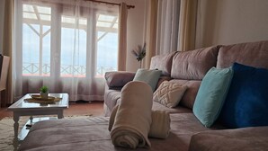 A cozy living room with a sofa, a coffee table, and a view through large windows. - Maiata Beach House 1 (Machico)