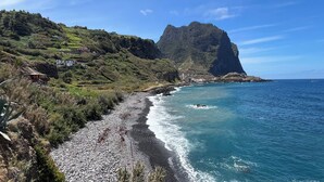 A coastal landscape with a rocky beach, lush greenery, and a prominent mountain in the background. - Maiata Beach House 1 (Machico)