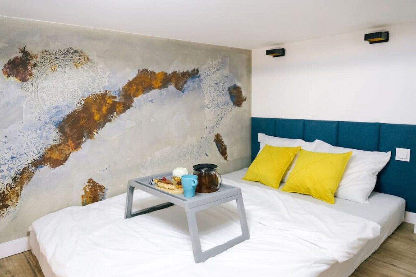 A bedroom with a bed, a bedside table with a mug, and a wall with a textured mural.
