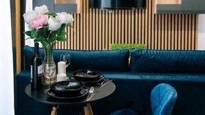 A modern living area with a blue sofa, a small round table with a vase of flowers, and a mounted TV on a wooden panel wall. - Łódź City Center Studio - Entresol & Balcony - by Rentujemy (Lódz)