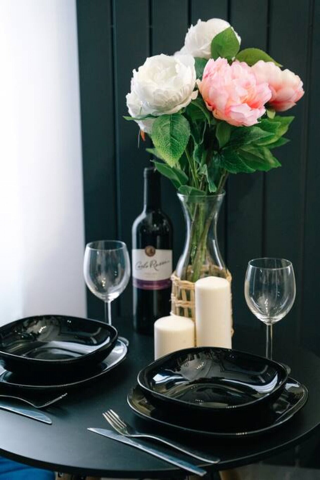 A table set with black plates, two wine glasses, a vase with flowers, a candle, and a bottle of wine.