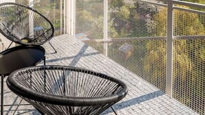 Ein Balkon mit zwei schwarzen Stühlen aus Draht und einem Metalltisch. Der Balkon verfügt über ein Metallgeländer und bietet Blick auf eine Stadtlandschaft mit Bäumen und Gebäuden.