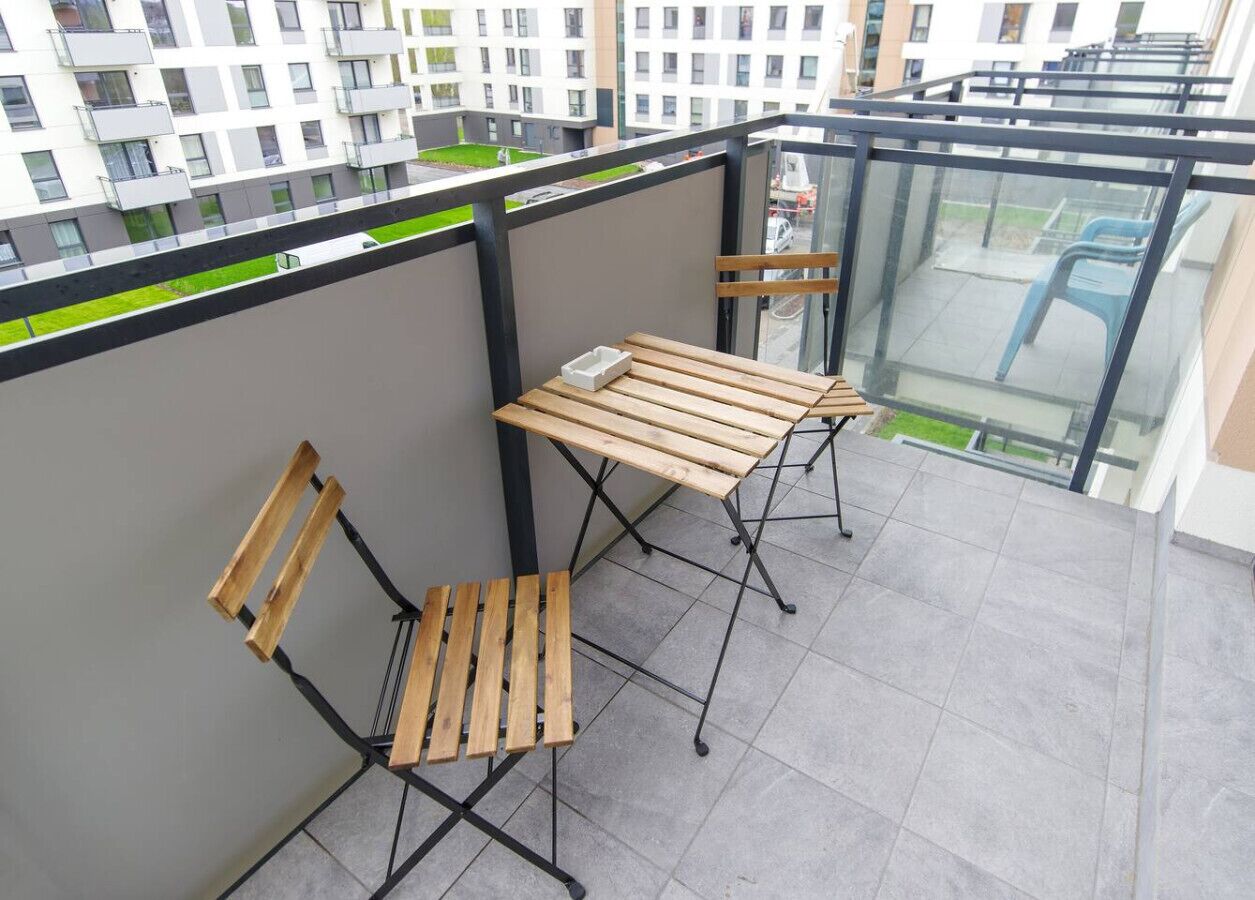 Ein Balkon mit zwei Holztischen und Stühlen, einer Glastrennwand und Blick auf eine Wohngegend.