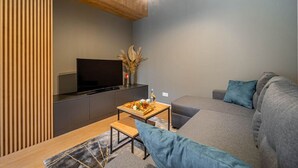 A modern living room with a flat-screen TV, a wooden table with a tray of snacks, a grey sofa, and a bench.
