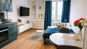 A modern bedroom with a bed, a desk, a chair, a TV, a shelf with plants, and a window with curtains. - Warsaw Studio Apartment - City View, Parks nearby by Rentujemy (Warszawa)